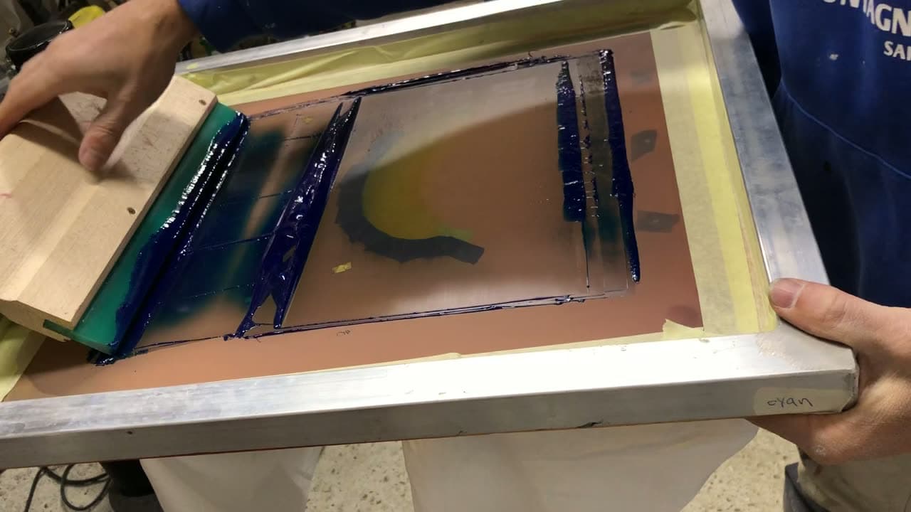Écran de sérigraphie encré de bleu et de vert, raclette en main, sur la table de l'atelier